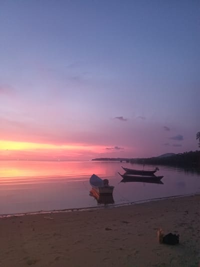 The image depicts a serene sunset over a body of water, likely the sea, in Baan Wok Tum, Thailand. The sky transitions from a light blue at the top to a vibrant mix of pinks, oranges, and yellows near the horizon, which reflects on the calm water surface. Three boats are visible: one close to the shore, seemingly beached, and two others further out, floating on the water. The shoreline is sandy, with some debris and vegetation visible. The distant shoreline shows signs of habitation and possibly a mountain range.