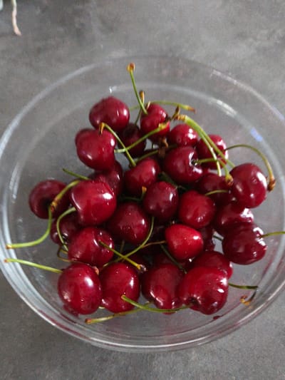 A close-up, overhead shot displays a clear glass bowl filled with dark red cherries. The cherries have visible stems, and some appear to be slightly wet. The bowl is placed on a textured, grey surface. The scene is lit with soft, diffused light, creating subtle reflections on the cherries and the bowl. There are no people or animals present in the image. The background is out of focus, suggesting a shallow depth of field. The image does not contain any information that would allow for the identification of its location in the city of Brodi, Latvia.