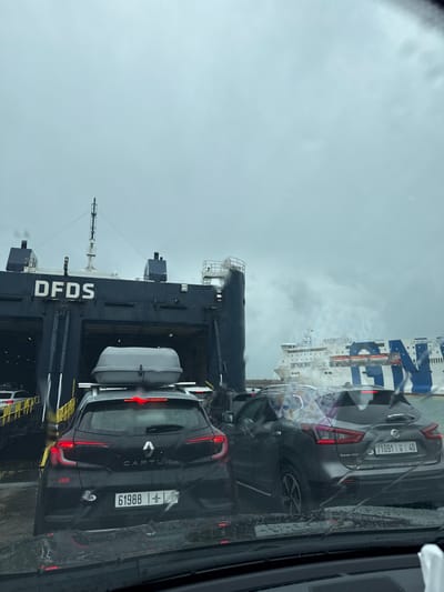 The image depicts a queue of vehicles waiting to board ferries at Tanger Med Port, near Ksar El Majaz, Morocco, under an overcast sky with visible raindrops on the viewer's windshield.
In the immediate foreground, from an interior vehicle perspective, raindrops are evident on the windshield. Directly ahead is the rear of a dark-colored Renault Captur SUV, equipped with a grey roof cargo box. Its Moroccan license plate reads "61988 ا 1". To the right of the Renault is the rear of a dark-colored Nissan Qashqai SUV, displaying the Moroccan license plate "71091 ا 40". Both vehicles appear stationary, positioned in a line to board a ferry.
Dominating the left and center of the scene is the stern of a large dark-blue roll-on/roll-off (Ro-Ro) ferry belonging to DFDS, with "DFDS" clearly visible in white lettering. Its vehicle ramp is lowered and open, revealing other vehicles positioned inside, indicating active boarding. Further in the background, to the right, a second large white and blue ferry from Grandi Navi Veloci (GNV) is visible, with "GNV" and "EXCELLENT" clearly printed on its hull. This GNV ferry appears to be either docked or maneuvering in the wider port area.