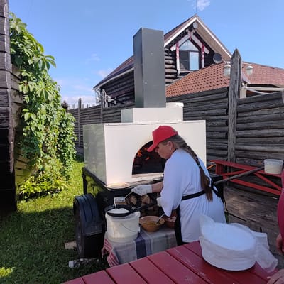 In Kudrino, Russia, a woman wearing a red cap and white apron is standing next to a white outdoor oven with a fire inside. She is wearing white gloves and holding a ladle, appears to be cooking. In front of her are various cooking utensils and ingredients, including a bucket of batter and a bowl of something yellow. The scene is set in a rustic outdoor area with a wooden fence and a log house in the background. The ground is covered in grass.