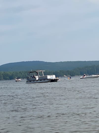 The image shows a lake with several boats. In the foreground, there is a large pontoon boat with people on board. To the left of the pontoon boat, there is a smaller boat. In the middle ground, there is a person on a paddleboard. To the right of the pontoon boat and the paddleboarder, there is a speed boat with people in it. In the background, there are trees and a hill. The sky is overcast. The scene is likely taking place in Duhamel, Canada, given the context.