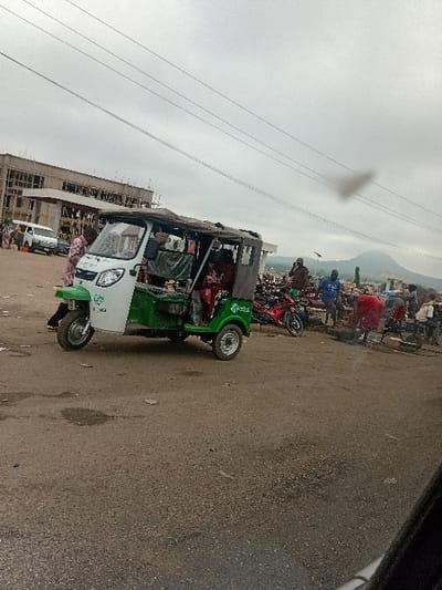 This image captures a busy, overcast daytime street scene in Itobe, Nigeria. The foreground consists of an unpaved or heavily worn asphalt road with visible dirt and minor debris. A two-tone green and white motorized tricycle, commonly known as a Keke Napep, is positioned slightly left of center, appearing stationary or in slow motion. It features a dark tarpaulin roof, and at least one individual dressed in red clothing is visible inside. Another person in light-colored patterned attire stands near the tricycle's front left wheel.
To the left background, a large, multi-story concrete building under construction is visible, characterized by exposed rebar and an unfinished facade. A white van is partially seen near this building. The mid-ground to the right features a cluster of approximately 5-7 individuals gathered around what appears to be market stalls or goods. One person in a red shirt is stooping over, while another in a blue shirt stands upright. A red motorcycle is parked behind this group, and a handcart or bicycle is also discernible. Distant, hazy hills or mountains are visible on the far right horizon under a cloudy sky. Three overhead power lines traverse the upper portion of the frame, with a small, dark, irregular smudge present in the upper sky, possibly a lens artifact or smoke.