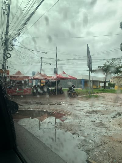 A street scene in Camaragibe, Brazil, is depicted from inside a vehicle, viewed through a rain-streaked windshield. The sky is overcast and grey. The foreground consists of an unpaved, muddy ground with numerous puddles reflecting the sky and surrounding elements.
In the midground, three red-orange pop-up tents are aligned. Under the leftmost tent, a banner partially reads "Rodoviária de Caldo de Cana." Several individuals are seated on chairs beneath these tents. To the right of the tents, two motorcycles are parked, with a light-brown dog standing near the front wheel of one. The ground in this area is rough, uneven, and saturated with water.
In the background, a complex network of overhead utility wires is visible, extending from multiple utility poles. Behind the tents, a building with a light-colored facade, featuring blue and yellow sections, is present. A large, dark, rectangular sign board, facing away from the viewer, stands on the right. Trees and green vegetation are visible on the far right.