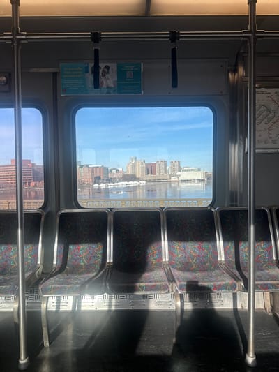 Inside a train car, rows of empty seats line up next to a window. The seats have a dark, patterned upholstery. Light streams in through the window, illuminating the seats with bright patches. Outside the window, a cityscape is visible, with buildings reflected in the water. A handrail runs along the length of the seats. Advertisements are placed on the wall between the windows.