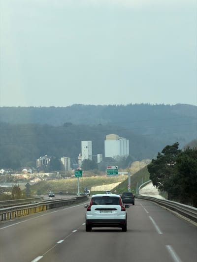 The image depicts an eastbound view from a multi-lane highway, identified as the A4 or N4, leading towards Pagny-sur-Meuse, France. The sky is uniformly overcast, casting diffuse light across the scene.
In the foreground, a three-lane asphalt highway is visible, marked with solid and dashed white lane lines. A white Volvo SUV, displaying French license plate GR-558-VV, occupies the leftmost visible lane, proceeding away from the viewer. Immediately ahead in the adjacent right lane, a dark-colored SUV is observed. Several other vehicles are discernible further down the highway in the distance. Steel guardrails line both sides of the roadway, with a white kilometer marker post labeled "28.4" affixed to the left guardrail. A circular speed limit sign, indicating "90" km/h, is positioned on the right shoulder.
In the midground, two large green highway directional signs are prominent. The sign on the left indicates routes for "A4 PARIS REIMS TROYES ST-DIZIER" with a left-pointing arrow. The sign on the right indicates "N135 NEUFCHÂTEAU BAR le DUC LIFFOL le BARROIS" with a right-pointing arrow. Multiple horizontal power lines stretch across the mid-distance.
The background reveals an industrial complex situated on a slight elevation to the left of the highway, comprising several large white structures, including tall silos and multi-story rectangular buildings, consistent with the Soufflet Agro Pagny-sur-Meuse site or similar industrial facilities in the town. Below and to the left of this complex, a residential area with multiple houses is visible. The entire distant horizon is characterized by densely wooded hills, primarily featuring bare deciduous trees and some conifers, appearing muted and hazy under the overcast conditions. No individuals are identifiable within the scene.