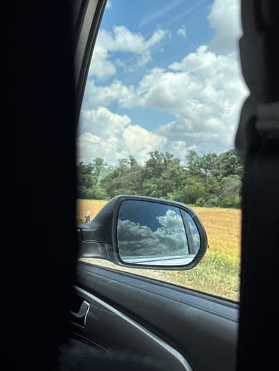 The image shows a view through a car window, looking at a side-view mirror. The mirror reflects a bright blue sky filled with fluffy white clouds. To the left of the mirror, a large portion of the sky and clouds are visible through the car window. Behind the mirror, the reflection shows trees and a field of dry, yellow grass or crops, suggesting a rural or agricultural setting. The car's interior, including part of the door panel with a chrome accent, is visible on the right side of the frame. The lighting indicates it is daytime, likely sunny, with clear skies. The setting appears to be Grachyovka, Russia, as indicated by the provided location context. There is no visible text in the image. The overall impression is a snapshot of a scenic drive on a pleasant day.