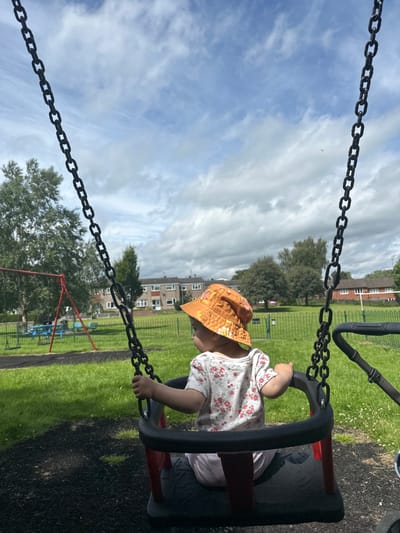A young child wearing a floral-patterned shirt and an orange bucket hat sits in a swing at a park. The swing has black chains and a black seat with red accents. The child is facing away from the camera, looking to their left. In the background, there is a grassy area with trees and residential buildings. The sky is cloudy. A red swing set is visible to the left of the frame. The ground beneath the swing appears to be dark, cushioned surfacing. The image was taken in a park in Macclesfield, United Kingdom.