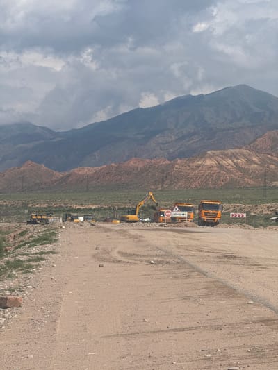 The image depicts a construction site in a mountainous, arid region, likely near Tong, Kyrgyzstan. In the foreground, a dirt road is visible with tire tracks. To the left of the road, there is a rocky embankment with some sparse green vegetation.

Further back, a construction area is active. Several yellow construction vehicles, including excavators and what appear to be loaders or bulldozers, are parked or in operation. Two large orange dump trucks are prominently positioned in the middle ground.

Traffic signs are present, including a red circular sign with a horizontal bar (indicating "no entry" or "closed") and a sign with an arrow pointing right and the word "объезд" (meaning "detour" or "bypass") in Cyrillic script.

In the background, a range of mountains dominates the landscape. The lower slopes of these mountains are characterized by reddish-brown, eroded rock formations and green vegetation. The upper mountains are covered in dark, possibly forested, slopes, and the sky is filled with heavy, overcast clouds. The overall impression is one of ongoing construction work in a natural, rugged environment.