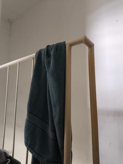 A teal colored towel is draped over a light-colored metal railing, possibly a staircase railing. The towel has stripes towards the bottom. The railing is made of vertical bars and a horizontal top bar. The wall in the background is a light beige color and appears to have some subtle staining. The lighting suggests an indoor environment, possibly with natural light from a nearby window. There is no indication of the location being in General Trias, Philippines.
