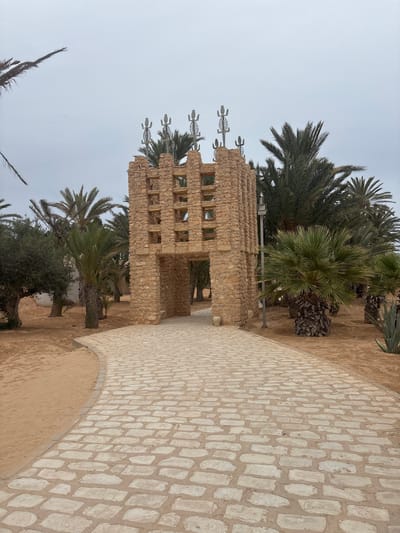 The image shows a stone archway with a textured, open lattice design, topped with decorative metal figures. The archway is constructed from rough-hewn, light-brown stones. A pathway made of light-colored cobblestones curves towards the archway. The pathway is bordered by sand and vegetation, including various types of palm trees and some spiky plants. The scene is set against an overcast sky. The archway appears to be a gateway to a park or an attraction within Tezdaine, Tunisia, a city known for its historical sites and natural beauty.
