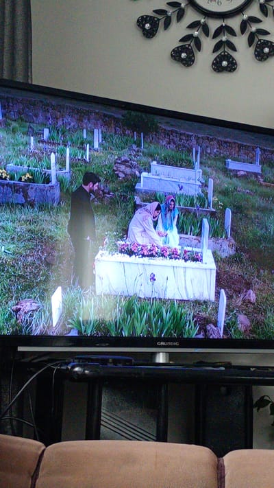 This image, reportedly taken in the city of Koniovets, Bulgaria, captures an indoor setting focused on a television displaying an outdoor scene.

In the foreground, the upper back and cushions of a light brown upholstered couch are visible. Immediately behind the couch, a black television stand supports a flat-screen television. A tangle of black cables and a small black electronic device are situated on the stand behind the television's lower bezel.

The television screen, bearing the "GRUNDIG" brand logo on its bottom right, displays an outdoor scene depicting a cemetery. Numerous white rectangular gravestones and grave enclosures are scattered across a grassy, gently sloping terrain. Some graves are surrounded by green leafy plants, while others are topped with vibrant purple and pink flowers. A prominent white grave enclosure with flowers is in the lower-mid section of the television screen, with a single purple flower in the immediate foreground of the TV scene. A low wall constructed of rough dark stones runs across the mid-section of the TV screen, defining the upper boundary of the cemetery area.

Within the television scene, three individuals are present near the prominent white grave. A man, dressed in a dark suit with a beard, stands on the left, looking downwards towards the grave. To his right, two women are kneeling or squatting beside the grave. The woman closer to the grave wears a light-colored (possibly beige or light brown) coat and headscarf, with her hands extended towards the flowers on the grave. The second woman, positioned behind the first, wears a light blue garment and headscarf, also looking towards the grave.

Above the television, a light beige wall is visible, adorned with a decorative black wall clock featuring ornate leaf and heart-shaped elements, some embellished with small shiny accents. A dark curtain or drape is partially visible along the left side of the wall.