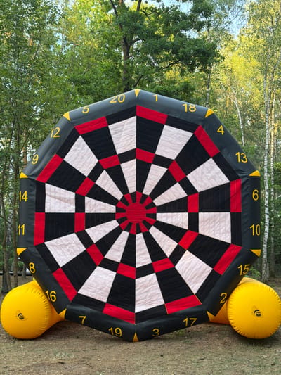 An inflatable dartboard with yellow numbers and red, black, and white sections stands outdoors in front of a forest of trees. The dartboard is supported by two large yellow inflatable balls at its base. The scene appears to be in a grassy area with some fallen leaves on the ground. The lighting suggests it is daytime, with sunlight filtering through the trees. The specific location is Ракітніцкі сельскі Савет, Belarus.