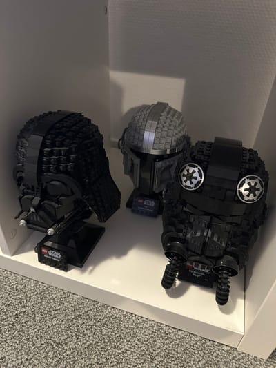 The image displays three LEGO Star Wars helmet models arranged on a white shelf within an indoor setting in Wanneperveen, Netherlands. From left to right, the models are a black Darth Vader helmet, a grey Mandalorian helmet, and a black TIE Fighter Pilot helmet. Each helmet rests on a black display stand that includes a "LEGO STAR WARS" plaque identifying the character. The Darth Vader helmet faces right, the Mandalorian helmet faces slightly right, and the TIE Fighter Pilot helmet faces forward. The shelf is white, and the background consists of a white wall or cabinet interior. The bottom left quadrant of the image shows a grey carpeted floor. No individuals or active interactions are present in the scene.