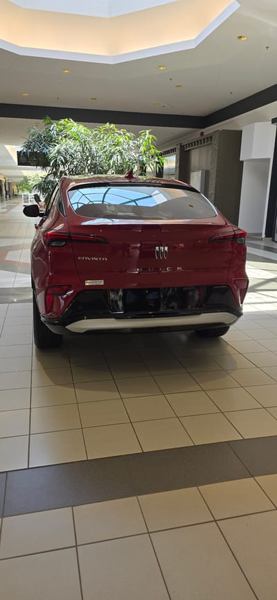 This is an image of the rear of a red Buick Envista car, parked indoors. The car is positioned at an angle, with its taillights and "BUICK" and "ENVISTA" badging visible. The interior of the car's rear window reflects the surrounding environment, including what appears to be a ceiling with skylights and lighting fixtures. A large indoor plant with lush green foliage is situated to the left of the car, partially obscuring its side. The floor is tiled with a pattern of light-colored squares and a darker gray strip. The lighting suggests it is daytime, and the indoor setting indicates a showroom, mall, or similar commercial space. No people are visible in the image. The visible text includes "BUICK", "ENVISTA", and a small sticker with text that appears to read "PU501-BMTHO". The overall impression is one of a clean, modern automotive display.