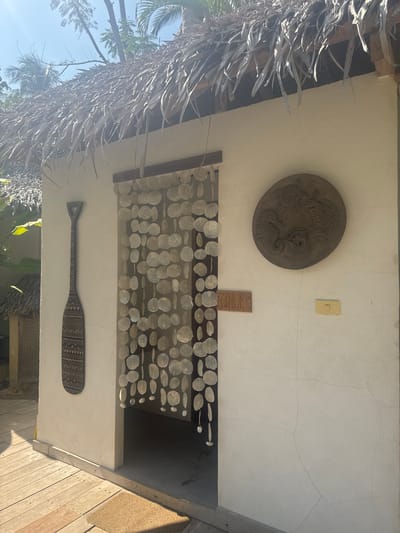 The image shows the exterior of a building in El Nido, Philippines. The building has a thatched roof with a circular decorative plate depicting marine life. There's an electrical switch plate above a rectangular plate with what looks like a design. A curtain of small white discs hangs below. A paddle decorated with geometric patterns is mounted horizontally below the curtain. The building appears to be constructed of wood and is exposed to sunlight. There are trees and vegetation visible in the background.