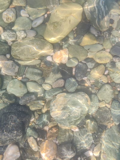 The image shows a collection of smooth, wet stones submerged in clear water. The stones vary in size, shape, and color, ranging from light grey and white to green, brown, and black. Sunlight reflects off the water's surface, creating dappled patterns and highlights on the stones. The scene depicts a natural environment, likely a riverbed or shoreline. There are no people or discernible actions taking place. The location is specified as Δημοτική Ενότητα Ευρυμενών, Greece, suggesting it is a place within that administrative division.