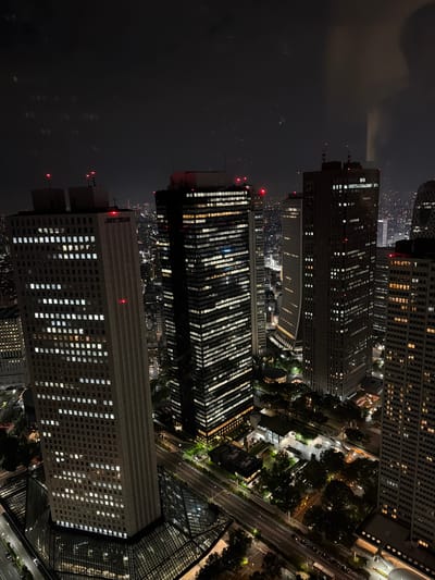 This image captures a nighttime aerial view of the Shinjuku area in Tokyo, Japan, focusing on a cluster of illuminated skyscrapers. The foreground is dominated by the Tokyo Metropolitan Government Building, with its distinctive twin towers and extensive glass atrium at the base. The building is lit up, with rows of windows glowing white, and red warning lights are visible on its upper sections. To the right of the Tokyo Metropolitan Government Building, other skyscrapers rise, also illuminated with countless windows. The scene shows a dense urban environment with streets visible below, lined with traffic lights and the headlights of moving vehicles. Green spaces and smaller structures are interspersed among the larger buildings. The overall impression is of a vibrant, bustling metropolis at night, with the architecture of Shinjuku prominently showcased.