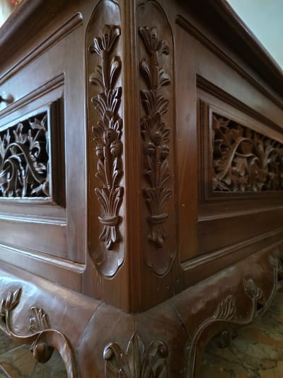 The image shows a detailed close-up of carved wooden furniture, likely a table or cabinet, with intricate floral motifs. The wood is a dark, rich brown and appears to be polished. The carvings are deep and elaborate, featuring stylized flowers and leaves. The furniture has a prominent corner with detailed decorative elements. The background is out of focus, but suggests an indoor setting with a marble or stone floor. There are no people visible in the image. The scene is static and focuses solely on the craftsmanship of the wooden object. There is no information or context provided within the image to suggest a specific location in Bireuenmeunasahblang, Indonesia.