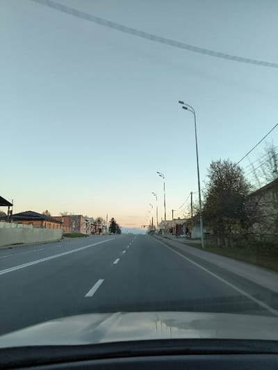 A straight asphalt road stretches into the distance beneath a soft, pale sky. Double white lines divide the lanes, with intermittent white dashes marking the center. Buildings line the roadside, including some with terracotta roofs. Streetlights illuminate the scene. Power lines crisscross the sky. The view is likely from inside a car, with a portion of the dashboard visible at the bottom.