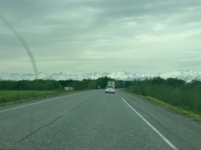The image depicts a cloudy, overcast day on a paved highway, likely in or near the city of Krasny, Russia, given the context. The highway stretches towards a distant mountain range capped with snow. Dense green trees line the sides of the road, with patches of green grass and some yellow wildflowers visible along the right shoulder. In the distance, a white SUV is driving away from the camera on the highway. There is a large truck also visible in the distance. On the left side of the road, there's a small, white and red building. On the right side of the road, there is a triangular warning sign with a black symbol on a white background, and a white sign with a black border and a black symbol. The sky is filled with thick, gray clouds. A faint, dark, funnel-shaped anomaly is visible in the upper left quadrant of the sky.