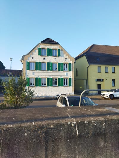 The image shows a multi-story building with a white stucco facade and a stepped gable, painted with the numbers "1677" across the upper portion. It has green shutters on its windows. To the right of this building is a yellow building with a brown tiled roof. In the foreground, a concrete wall runs horizontally, partially obscuring two cars parked on a street. A tree with green foliage is visible to the left of the white building. In the background, a cell tower can be seen. The scene appears to be a street in Leverkusen, Germany.