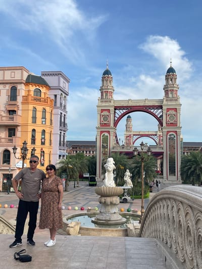 An elderly Asian couple, the woman in a floral dress and the man in a t-shirt and pants, pose for a photo on a stone bridge with intricate carvings. They stand on a patterned tiled plaza in front of a whimsical architectural structure resembling a red and white bridge with two towers. Behind them, colorful buildings with balconies and arched windows line the street, creating a picturesque, European-inspired urban setting. Palm trees are scattered throughout the plaza, and a fountain with sculptures is visible. The sky above is a clear blue with scattered white clouds. In the distance, other pedestrians and a small green vehicle are visible. The overall scene suggests a tourist destination or a themed outdoor space.