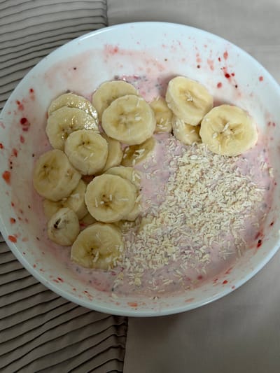 A top-down view shows a white ceramic bowl filled with a pink, possibly berry-flavored, oatmeal or porridge. Sliced bananas are arranged on one side of the bowl, partially submerged in the oatmeal. A pile of shredded coconut flakes sits on top of the oatmeal on the other side of the bowl. The bowl's rim has streaks and splatters of a reddish substance, possibly berry juice or a stain from the ingredients. The bowl is placed on a textured, gray surface. There is no information in the image that would pinpoint the location to Test Valley, United Kingdom. No people or actions are discernible.
