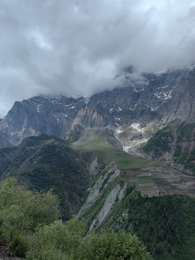 A landscape photograph captures a mountainous region under a cloudy sky. The towering mountains, some with snow patches, dominate the upper half of the frame, with jagged peaks and steep slopes. The lower half of the image shows a valley with terraced fields, a village, and lush green vegetation. The clouds are thick and gray, obscuring parts of the mountain peaks and creating a diffused light. The scene is one of natural beauty and human cultivation in a rugged environment. The location is identified as Gondhla, India.
