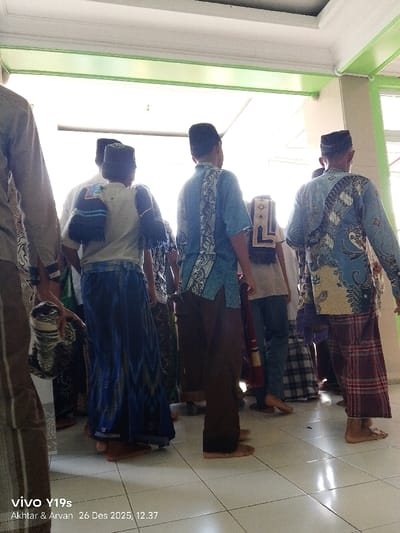 A group of barefoot men, likely Muslim, are standing together in an indoor space with tiled floors and light green trim. Most are wearing peci hats. Several have prayer rugs draped over their shoulders. Some are wearing patterned shirts and sarongs. The men appear to be gathered near a doorway or entrance. The date and time "26 Des 2025, 12:37" are visible at the bottom.