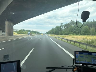 The image is taken from the perspective of someone driving on a highway in Germany, likely the A10 autobahn, given the presence of a navigation system displaying "Ludwigsfelde" and "Schönefeld, Berliner Ring." A highway overpass is visible in the foreground, casting a shadow over the lanes. Several vehicles, including trucks and cars, are on the road. The right side of the highway features a guardrail and a grassy verge with trees and vegetation. In the lower right corner, a GPS navigation device is mounted on the dashboard, showing a map with a route highlighted and indicating the destination "Ludwigsfelde" and current location "Schönefeld, Berliner Ring." The sky is cloudy.