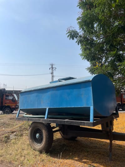 The image depicts a blue water tank mounted on a trailer with wheels, situated outdoors in what appears to be a rural or semi-urban area. The tank is cylindrical at one end and rectangular along its length. The trailer's frame is black. The trailer is parked on dry, grassy ground. In the background, power lines and a utility pole are visible. Also, a red truck is partially visible. The presence of the truck and utility pole suggests a location within or near a populated area. The city of Anajpur, India is presumed to be the location based on the prompt instructions. There are some green leaves on the top left corner.