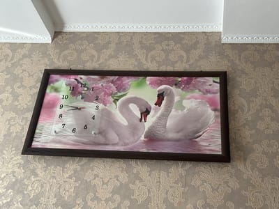 The image shows a framed clock with a picture of two swans and pink flowers. The clock has Arabic numerals and two hands pointing to approximately 1:55. The clock is hanging on a wall with wallpaper that has a floral pattern. The wall is also adorned with white crown molding. The scene appears to be an interior shot.