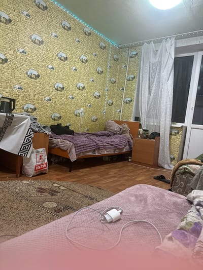 This is a cluttered bedroom in an apartment in Corlăteni, Moldova. A bed with purple bedding is the central focus, appearing unmade. To the left of the bed is a wooden nightstand with a bag from "Kaufland" on it, along with a black bag and what appears to be a kettle. The walls are covered in yellow wallpaper with a stone pattern and images of houses. A white sheer curtain hangs across a window on the right side of the room. The floor is covered with a patterned rug and hardwood flooring. A charger and cable are visible on a pink blanket in the foreground. There are no people visible in the image. The room appears to be lived-in and somewhat disorganized.
