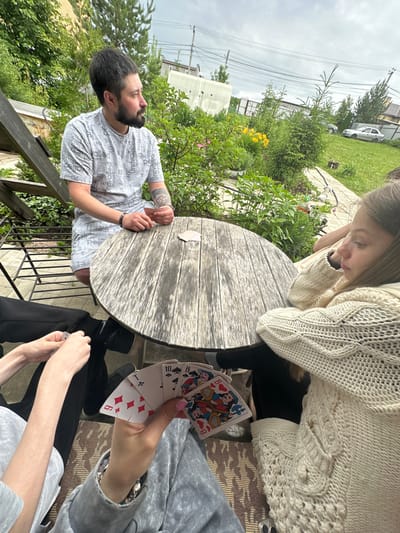 The image captures an outdoor card game in Bol'shaya Mos', Russia, under an overcast sky. A round, weathered wooden table sits centrally on a patterned rug, surrounded by three visible individuals.

To the left, a man with short dark hair and a trimmed beard, wearing a light grey t-shirt and shorts with repetitive text, is seated on a dark metal chair. A tattoo is visible on his right forearm. He has a small white object near his mouth and his hands rest on the table beside a single face-down playing card, as he looks to the right.

To the right, a woman with long, light brown hair wears a cream-colored, cable-knit sweater. Her forearm rests on the table as she gazes downwards and to her left. A light-colored smartphone case is visible on the rug near her side.

In the immediate foreground, a person's hands, wearing grey sweatpants and a matching long-sleeved top, hold a fan of five playing cards in their right hand: the 9 of Diamonds, 10 of Hearts, 10 of Spades, King of Clubs, and King of Hearts. Pink nail polish is visible on some fingernails, and a beaded bracelet adorns their left wrist. Adjacent to these, another pair of hands with fair skin, wearing black pants, manipulate a playing card showing a '9'.

The background consists of a lush garden with various green foliage, bushes, and trees, including a prominent conifer. Yellow flowers are visible within the greenery. Beyond the garden, a paved path leads to a grassy area where two light-colored sedan cars are parked. Distant residential structures and fencing are discernible, and multiple utility lines stretch across the upper sky.