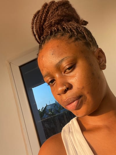 A person poses for a selfie indoors, illuminated by warm light. They have dark skin, brown hair styled in small twists piled atop their head, and facial piercings in their nose and ear. A white tank top is visible on their right shoulder. Behind them is a window with a partial view of blue sky and green foliage.
