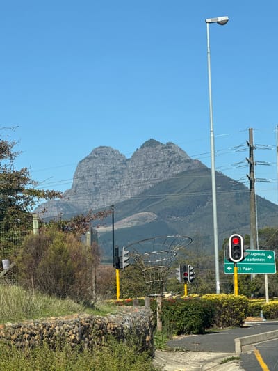 The image shows a road in Stellenbosch Local Municipality, South Africa. The road has traffic lights that are red. A green road sign points up to Klapmuts and Kraaifontein, and down to Paarl. In the background is a large mountain. A street lamp and power lines are visible. The side of the road has bushes and a stone wall.