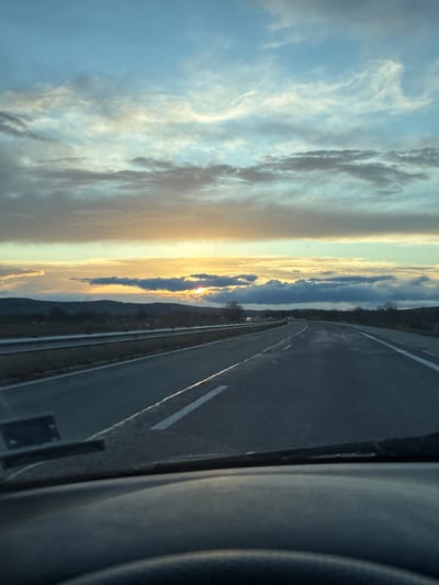 The image shows a highway road with painted white lines and barriers on the sides. The sky is visible in the background, featuring a sunset with clouds and a yellow glow. The scene is taken from inside a vehicle. The location is Rogozinovo, Bulgaria.