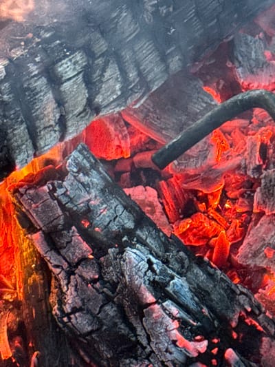 The image shows a close-up view of burning embers and charred wood. The embers glow with an intense red and orange light, illuminating the rough, textured surfaces of the blackened wood. The wood appears to be in various stages of combustion, with some pieces still retaining their original shape while others are crumbling into ash. The background is dark and out of focus, emphasizing the fiery core of the scene. There are no people or specific objects identifiable beyond the burning wood and embers. The image provides no information about the location in Imelevo, Russia, nor does it depict any specific actions or interactions.
