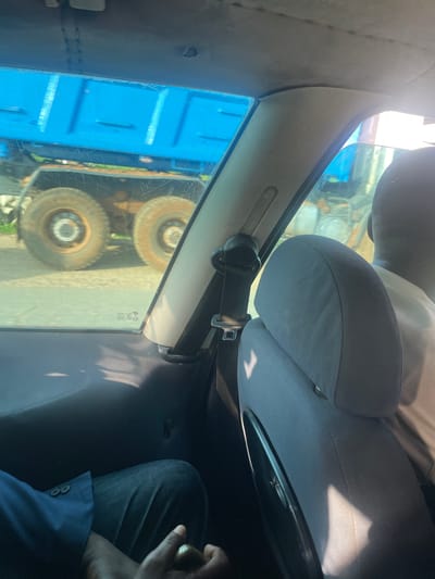 The image captures a view from inside a vehicle in Yola, Nigeria, looking outwards. A bright blue truck, likely a dump truck or similar construction vehicle, is the prominent subject outside the car, seen through the windows. The truck has large, weathered tires and its distinctive blue cargo bed is clearly visible, suggesting it is parked or moving slowly on a road. The environment outside appears to be a typical urban or semi-urban setting with hints of greenery. Inside the vehicle, the interior is partially visible. A person's head and shoulder, seen from behind, are in the foreground, suggesting they are a passenger. The seat upholstery is grey. A seatbelt mechanism is visible, attached to the car's frame. Another person's legs, wearing denim jeans and a blue jacket, are also visible in the lower left portion of the image, indicating at least two people are in the car. The lighting suggests it is daytime, with sunlight casting shadows and reflections on the interior surfaces and windows. The overall impression is a snapshot of daily life and movement within the city of Yola.
