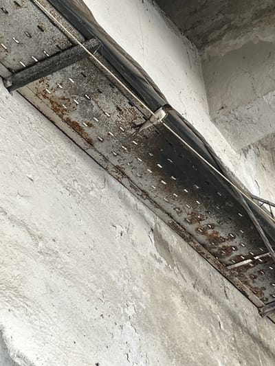 A small bird, likely a swallow or swift, is perched on a thin wire that runs parallel to a rusty, perforated metal tray. The tray is affixed to a rough, white-painted concrete wall, and appears to be part of an industrial or utility infrastructure. The bird is facing to the left and its feathers are a mix of dark and light brown. The wall shows signs of wear and tear, with patches of discoloration and peeling paint. Above the tray, dark conduits or pipes are visible, secured by metal clamps. The overall environment suggests an outdoor or semi-enclosed urban setting, possibly in an alleyway or under an overpass. The image was taken in Djerman, Bulgaria.