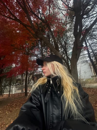 A woman with long blonde hair wears a black leather jacket and a black baseball cap with pink lettering. She stands outdoors among trees with reddish-brown leaves. A metal fence and some buildings are visible in the background, blurred by the focus on the woman and foliage in the foreground. The ground is covered with fallen leaves. The photo is shot from a low angle.