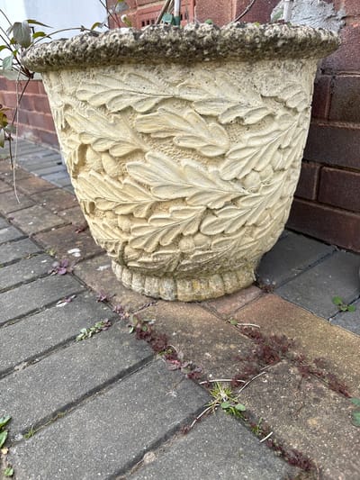 A weathered, ornate, light-colored stone planter sits on a paved surface in what appears to be an outdoor area. The planter is adorned with a raised leaf pattern. It is positioned near a brick wall. The paved surface consists of rectangular gray concrete pavers with weeds growing in the joints between the pavers. The location is implied to be Minsterley, United Kingdom.