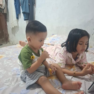 Two children are lying on a bed with a floral pattern. The child on the left, a boy, is wearing a green and white shirt and grey and white striped shorts. He is holding a yellow object in his hand and looking to his left with his mouth open. The child on the right, a girl, is wearing a pink pajama top with cartoon patterns. She is looking down at her hands, which are intertwined. A tablet is partially visible on the bed to her right. In the background, a wall with some items hanging on it is visible, including what appears to be clothing. The scene is likely indoors, possibly in a bedroom. The presence of floral patterns and a casual indoor setting suggests a domestic environment within the city of Reudeuep, Indonesia.