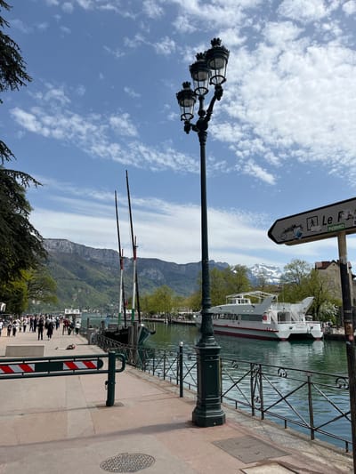 This is an outdoor scene at Annecy, France, on a partially cloudy day. The view is from a sidewalk or pedestrian area along a waterfront. To the left of the frame is a tall, ornate street lamp with multiple glass-enclosed lights. A directional sign pointing towards "Le F" is attached to another pole in the scene. In the background, a mountain range is visible, some of which has snow on it. A white boat is moored along the quay. The water of the canal or lake is rippled. The embankment is protected by metal railings, and there's a safety barrier with red and white stripes. A crowd of people is walking along the sidewalk near the trees.