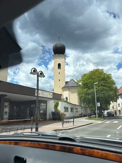The image captures a view from inside a vehicle, looking out onto a street in Reutte, Austria. A prominent church tower with a black onion dome and a clock face dominates the upper center of the frame. To the left of the tower, a building with a dark awning and a sign that reads "Brautmoden Christine" is visible. This indicates a bridal shop, and the sign also includes the words "Hochzeits- & Festmode" (Wedding & Evening Wear). In front of the shop, there are two black lampposts. A large, leafy green tree stands to the right of the church, and beyond it, residential buildings with pitched roofs are visible. The street in the foreground has a crosswalk and is being used by a black car and a motorcycle. The sky is cloudy, with patches of blue sky. The interior of the car, including the dashboard with wood trim and a portion of the windshield, is visible in the foreground.