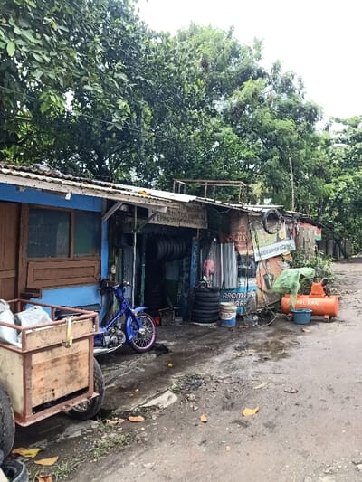 A small roadside tire repair shop nestles under a canopy of green trees. The structure, a mix of blue paint and corrugated metal, is cluttered with tires, tools, and miscellaneous items. A blue moped sits near the entrance, while an orange air compressor stands on the right. A wooden cart with tires leans into the scene, adding to the overall disarray. The ground is muddy and strewn with leaves.