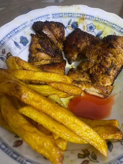 A close-up, high-angle shot captures a meal consisting of grilled chicken and french fries served on a patterned plate. The plate is white with a slightly scalloped edge, adorned with a blue border featuring a repetitive geometric design, and intermittent floral motifs in blue and yellow.
Two pieces of grilled chicken, seasoned with dark spices and showing some charring, are positioned on the left side of the plate. The chicken appears juicy and cooked through. To the right of the chicken, a small, deep red pool of sauce, likely ketchup or chili sauce, is present.
Occupying the right and central portions of the plate is a generous serving of golden-brown french fries. The fries appear crispy and are unevenly cut, suggesting they might be homemade or hand-cut. Some oily residue is visible on the plate, particularly around the food items.
The background is out of focus, suggesting the image was taken indoors, possibly on a table. No people are visible in the frame, and there is no discernible text. The lighting is soft but sufficient to highlight the textures and colors of the food.
