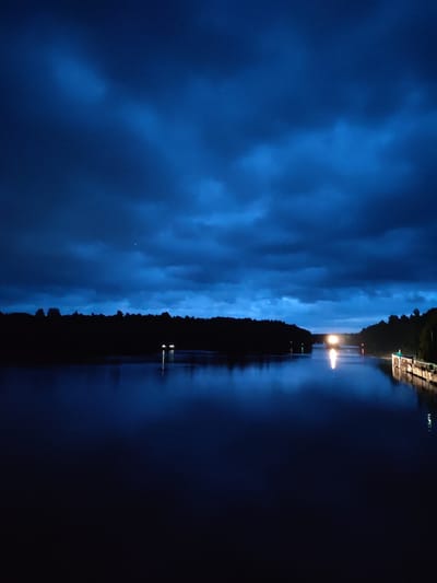 The image depicts a body of water, likely a river or canal, at dusk or night. The sky is dark blue with heavy cloud cover, creating a dramatic and moody atmosphere. On either side of the water, silhouetted against the sky, are dense tree lines. In the distance, a bright light emanates from what appears to be a vessel or structure on the water, casting a reflection. Along the right bank, some illuminated structures and lights are visible, possibly indicating a waterfront area or a lock system. There are no people visible in the image. The scene suggests a quiet, possibly remote location. The image is too dark to definitively place it in Devyatiny, Russia, however the dense forests and waterways are consistent with the general geography of that region.