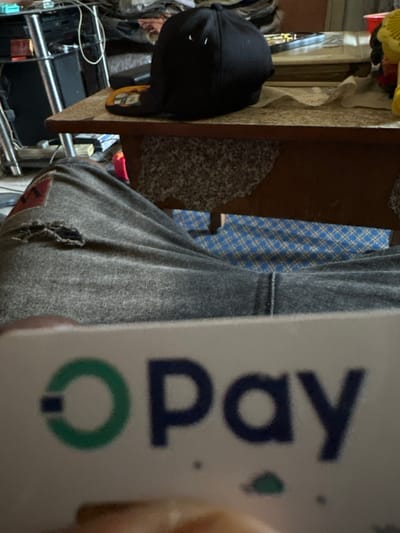 The scene is an indoor environment within Iriebe, Nigeria. The image captures a first-person perspective, primarily featuring the legs of a seated individual and a card held in their hand. The individual wears dark grey, distressed jeans with a prominent tear on the left knee area and a red patch on the upper left thigh. In the immediate foreground, a white card with the green and blue "O Pay" logo is held, partially obscuring the lower portion of the image; the text is slightly out of focus. Beyond the individual's legs, a light brown wooden table with a speckled light-colored surface is visible. A black baseball cap rests on the table, oriented with its brim to the left, displaying two white eyelets on its crown. To the right on the table, a yellow fan, a red cup, and other miscellaneous items are present. Below the table, the floor is covered by a blue and white checkered patterned fabric or mat. The background of the room behind the table appears cluttered with electronic devices and various household items.