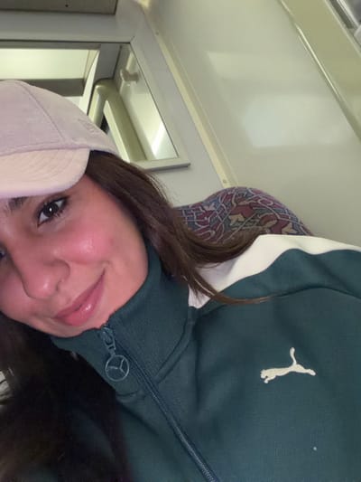 A woman with long brown hair and a light pink cap is looking at the camera. She is wearing a green Puma jacket with a white stripe on the shoulder. The backdrop seems to be inside a train with a patterned headrest and a white wall. A mirror and a luggage rack are visible in the background. The image is slightly rotated counterclockwise.